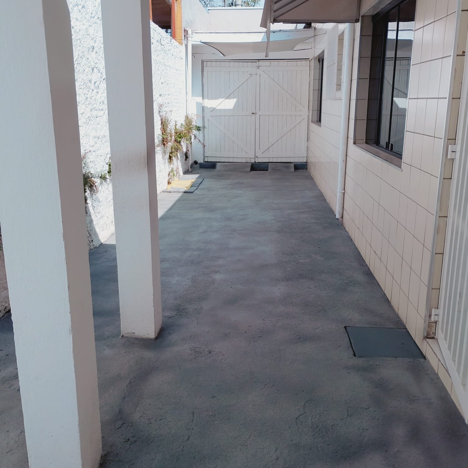 Acesso lateral coberto do imóvel, em concreto, com pilares, cobertura e portão branco de madeira ao fundo.