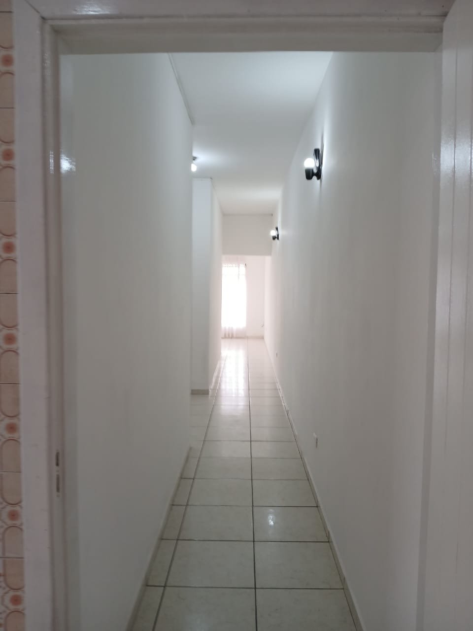 Corredor central interno do imóvel, com paredes brancas, arandelas de parede e piso claro.