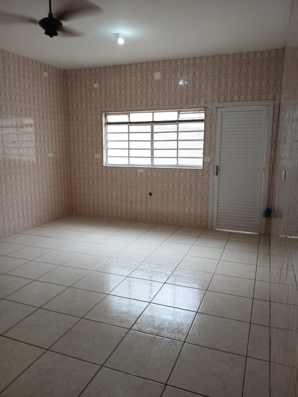 Ambiente interno amplo e vazio, com janela grande gradeada, ventilador de teto, azulejo original nas paredes e piso frio claro.