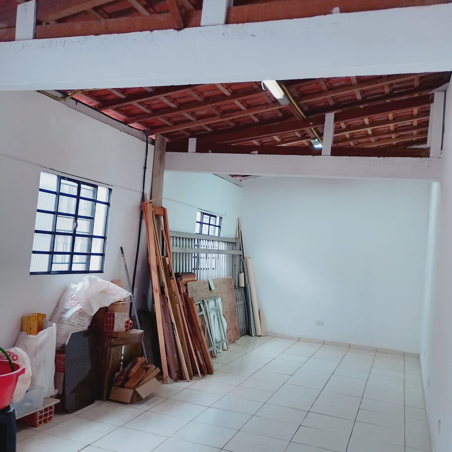 Salão interno amplo com pé-direito alto e estrutura de telhado em madeira aparente, piso branco e janela gradeada.