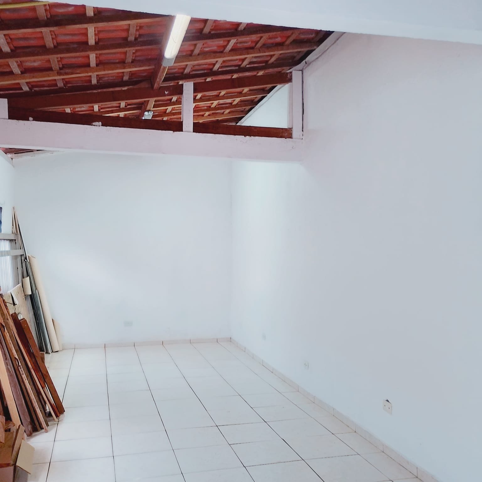 Segundo ângulo do salão interno vazio, com piso branco, paredes claras e telhado em madeira aparente.
